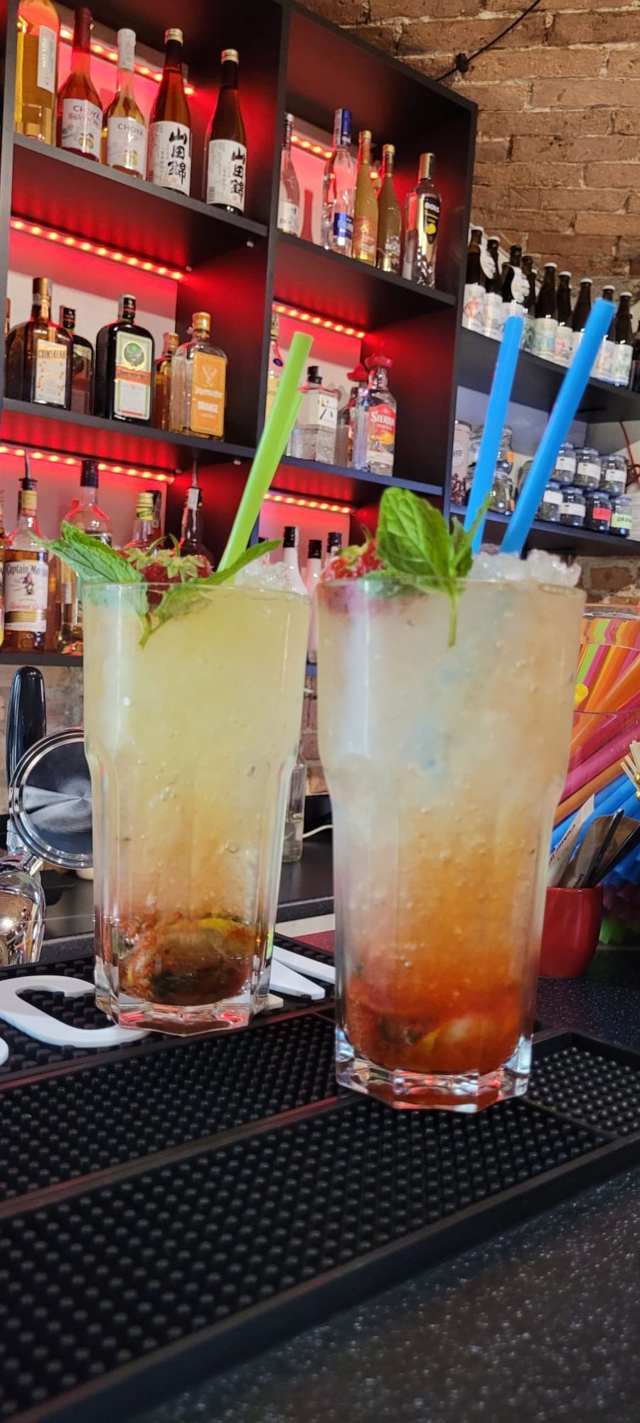 Dwa kolorowe koktajle owocowe na barze, z miętą i słomkami, w tle butelki alkoholu.