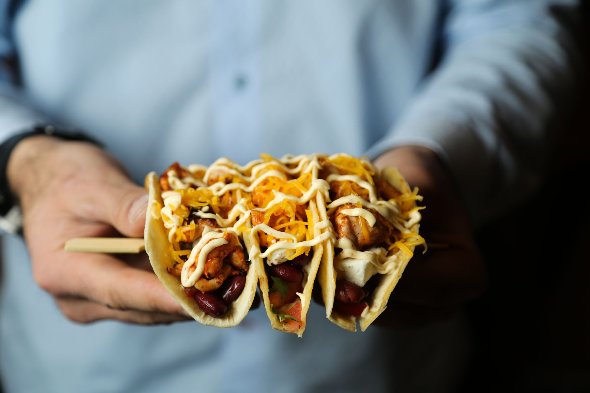 Trzy tacos w rękach, z kurczakiem, serem, warzywami i sosem, na tle niebieskiej koszuli.