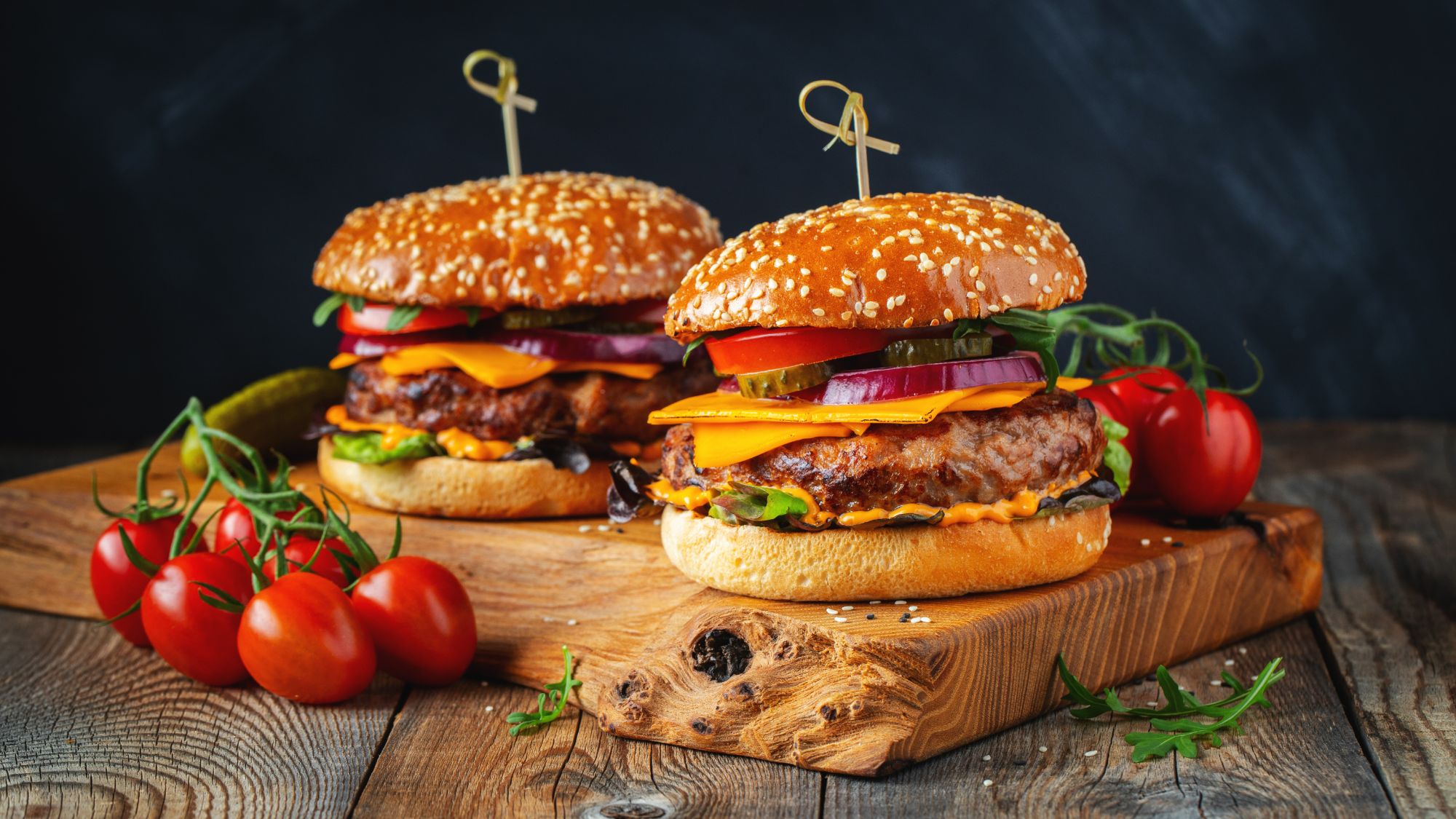Dwa soczyste burgery w bułkach sezamowych z warzywami na drewnianej desce, obok pomidory.