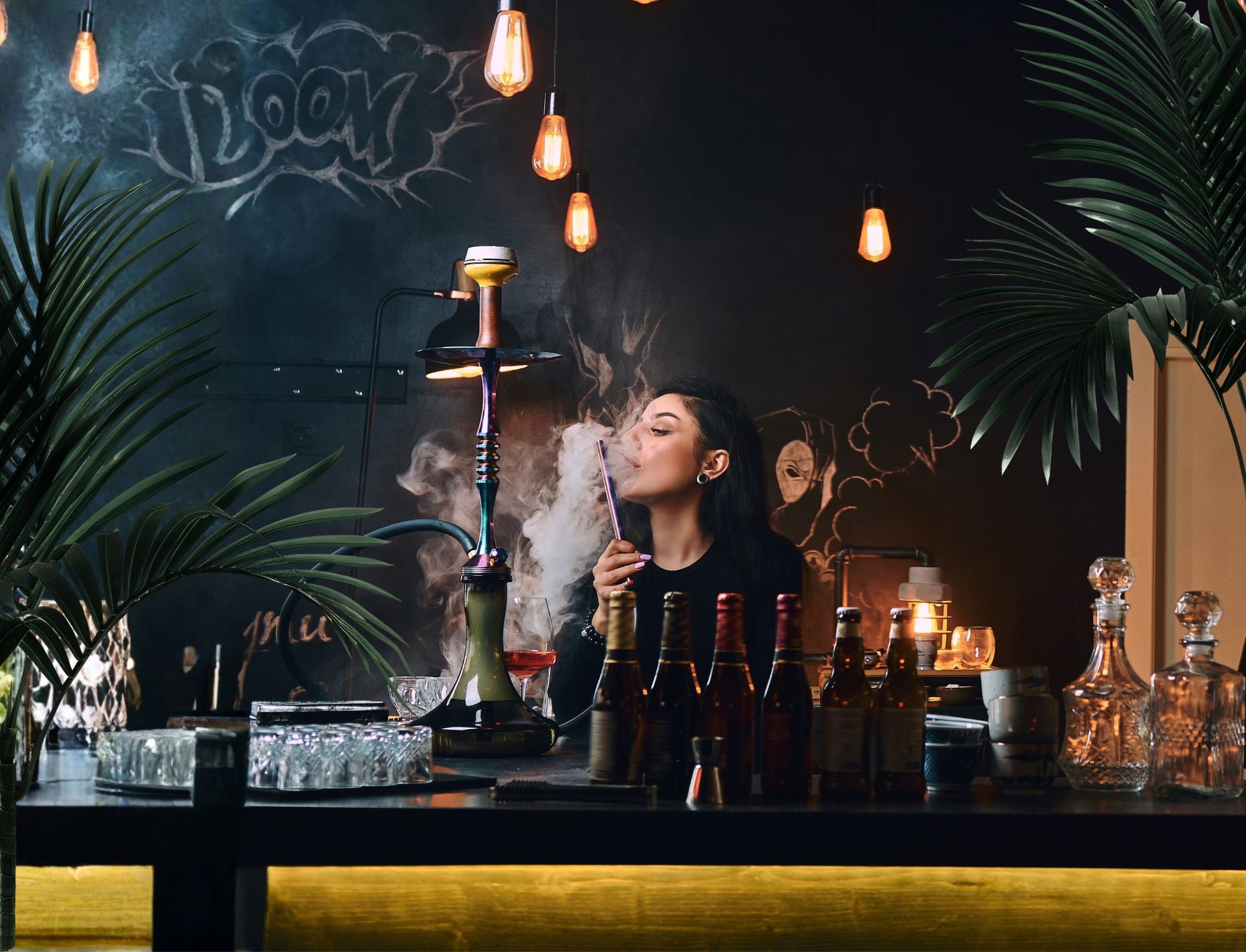 Kobieta w barze pali z fajki wodnej, wokół butelki alkoholu i wiszące lampy.