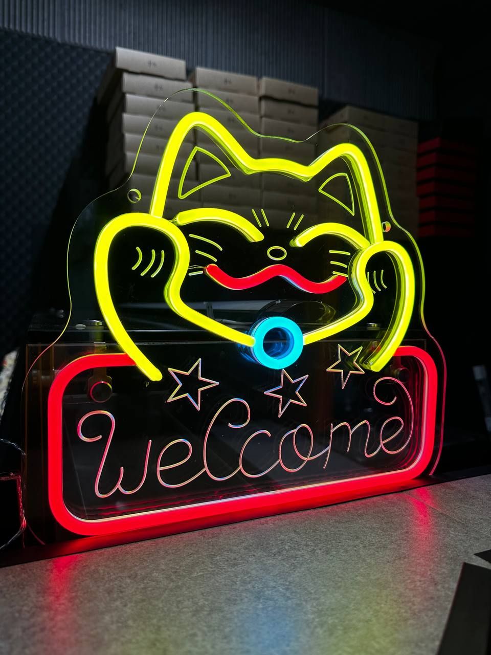 Neonowy szyld "welcome" z wizerunkiem uśmiechającego się kota z wąsami i gwiazdkami, emitujący kolorowe światło.