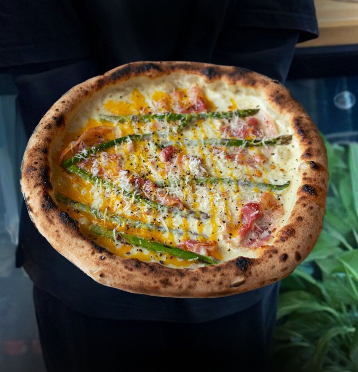 Pyszna pizza z serem, szparagami i boczkiem, trzymana przez osobę w czarnej koszulce.