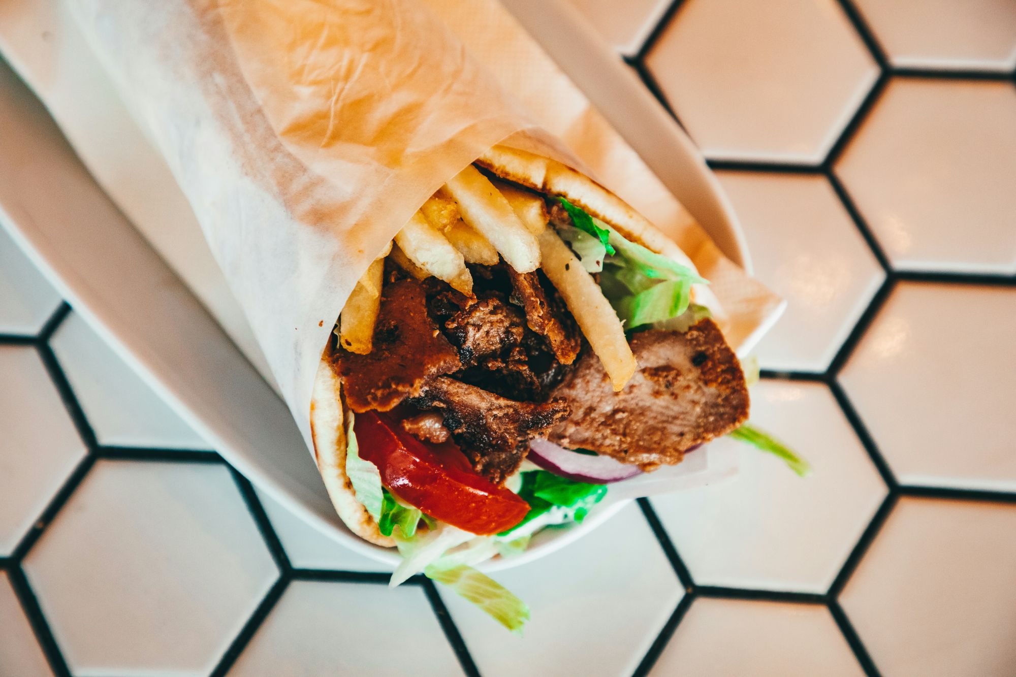 Burrito z mięsem, frytkami, sałatą, pomidorem i cebulą na talerzu, na tle biało-czarnych płytek.