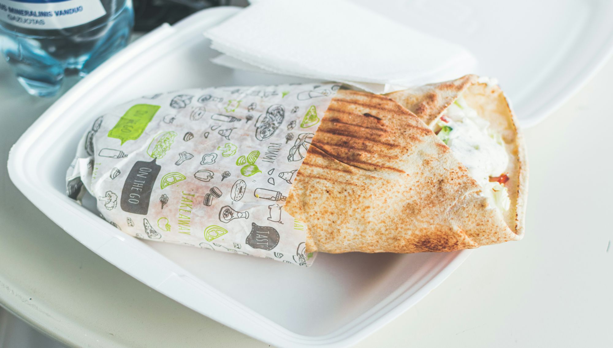 Kebab w cienkim cieście w opakowaniu na białym talerzu, obok butelka wody i serwetka.