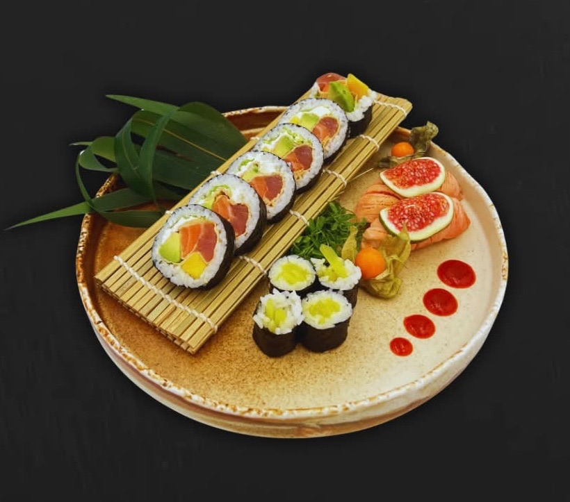 Talerz sushi z kolorowymi rolkami, wędzonym łososiem, figami i sosem na ciemnym tle.