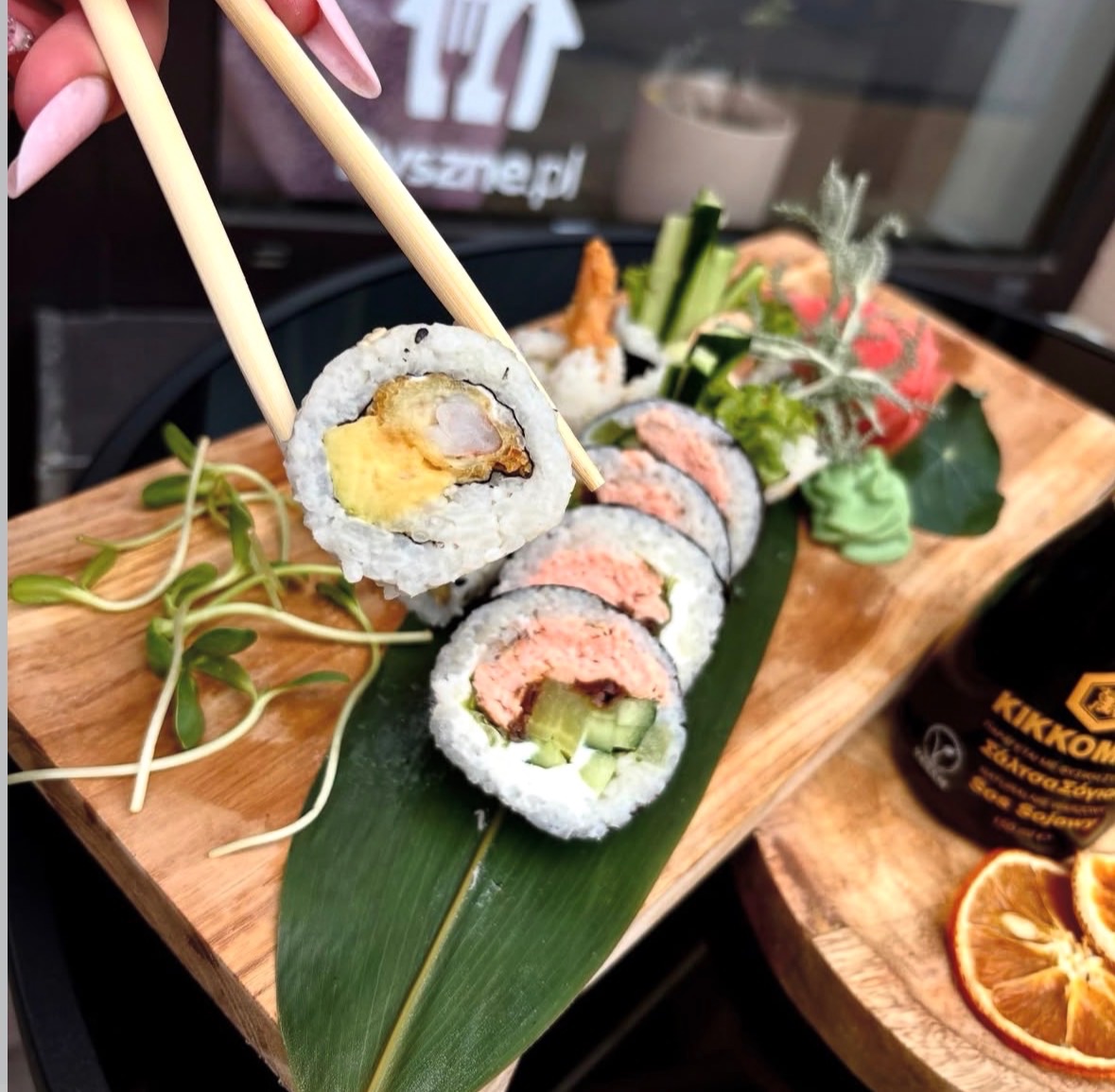 Sushi na drewnianej desce, podawane z pałeczkami, z różnorodnymi składnikami i sosem sojowym w tle.
