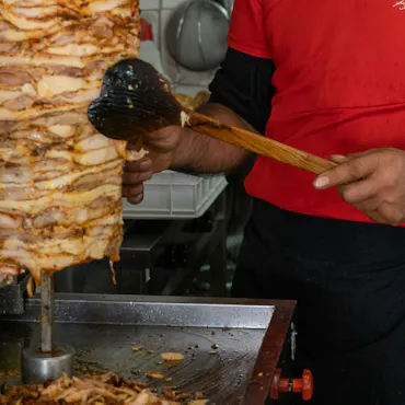 Mężczyzna w czerwonej koszulce trzymający łyżkę, przy rożnie mięsa w restauracji.