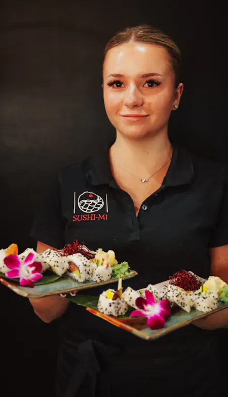 Kobieta w czarnej koszulce trzymająca dwa talerze z sushi ozdobione kwiatami.