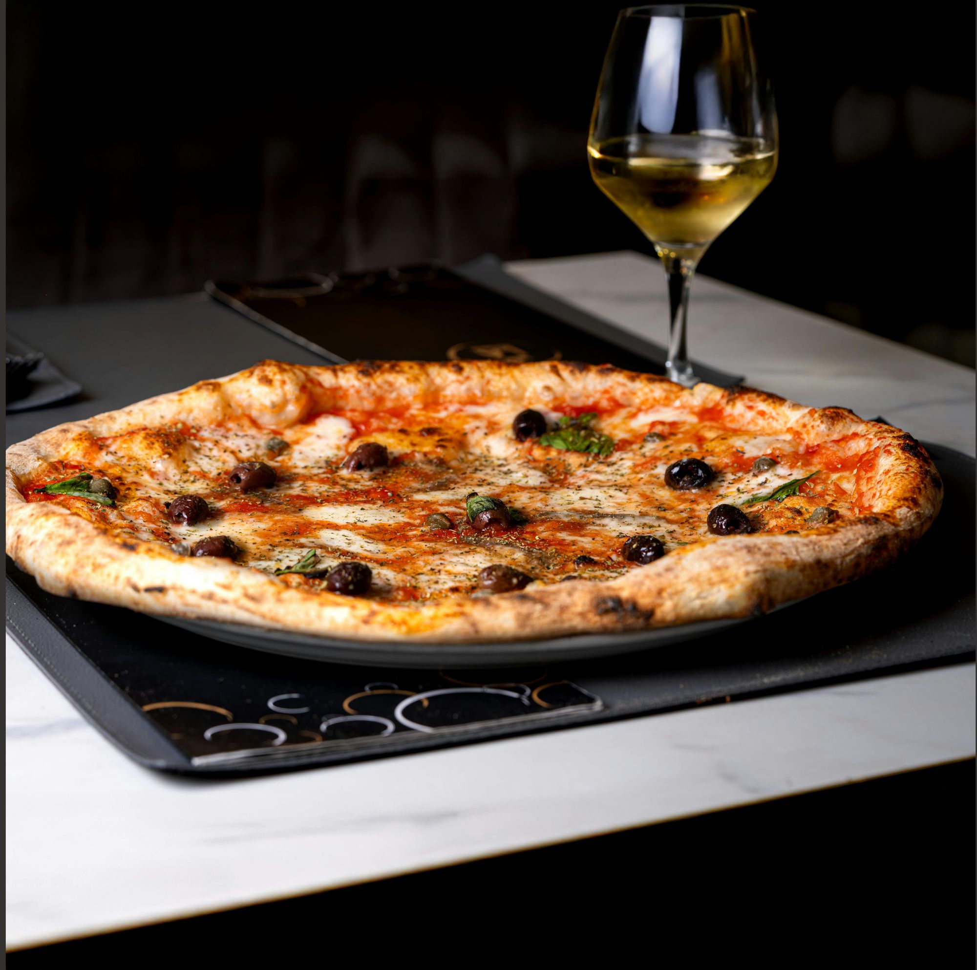 Pizza s olivami a bylinkami na čiernom podnosu, s pohárom bieleho vína na pozadí.