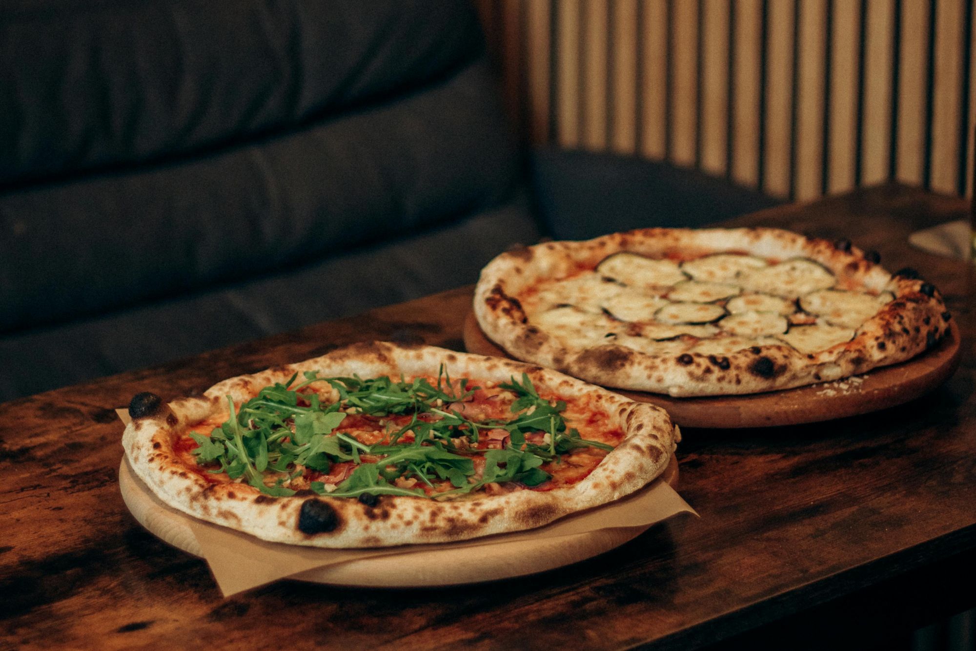 Dve pizze na drevenej doske, jedna s rajčinami a rukolou, druhá s cuketou a syrom, na stole v reštaurácii.