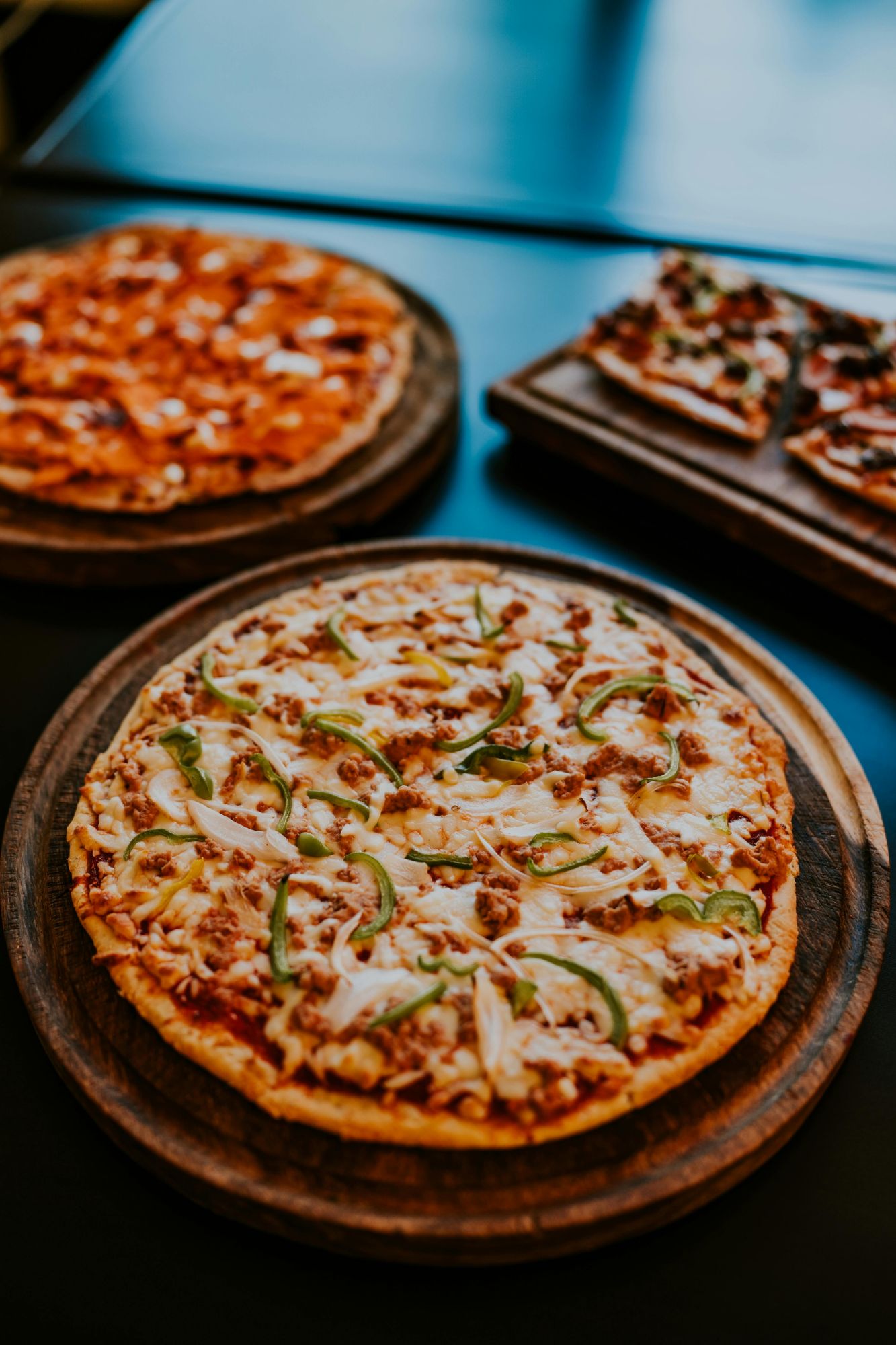 Trzy rodzaje pizzy na drewnianych talerzach: jedna z mięsem, cebulą i papryką, druga w różnych smakach.