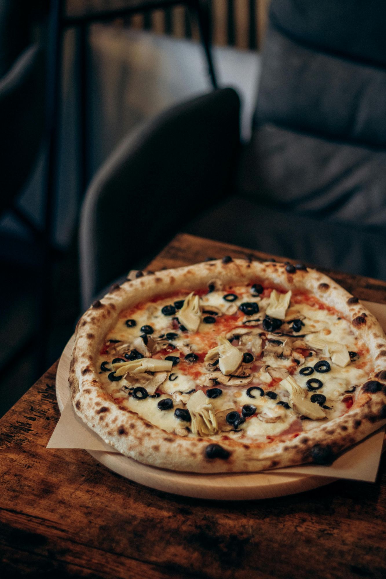 Pizza z oliwkami i karczochami na drewnianym talerzu, ustawiona na stole w restauracji.