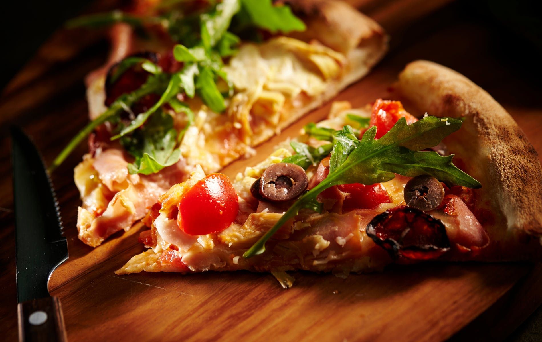 Plasterki pizzy na drewnianej desce, z różnymi dodatkami, takimi jak rukola, pomidory i oliwki.