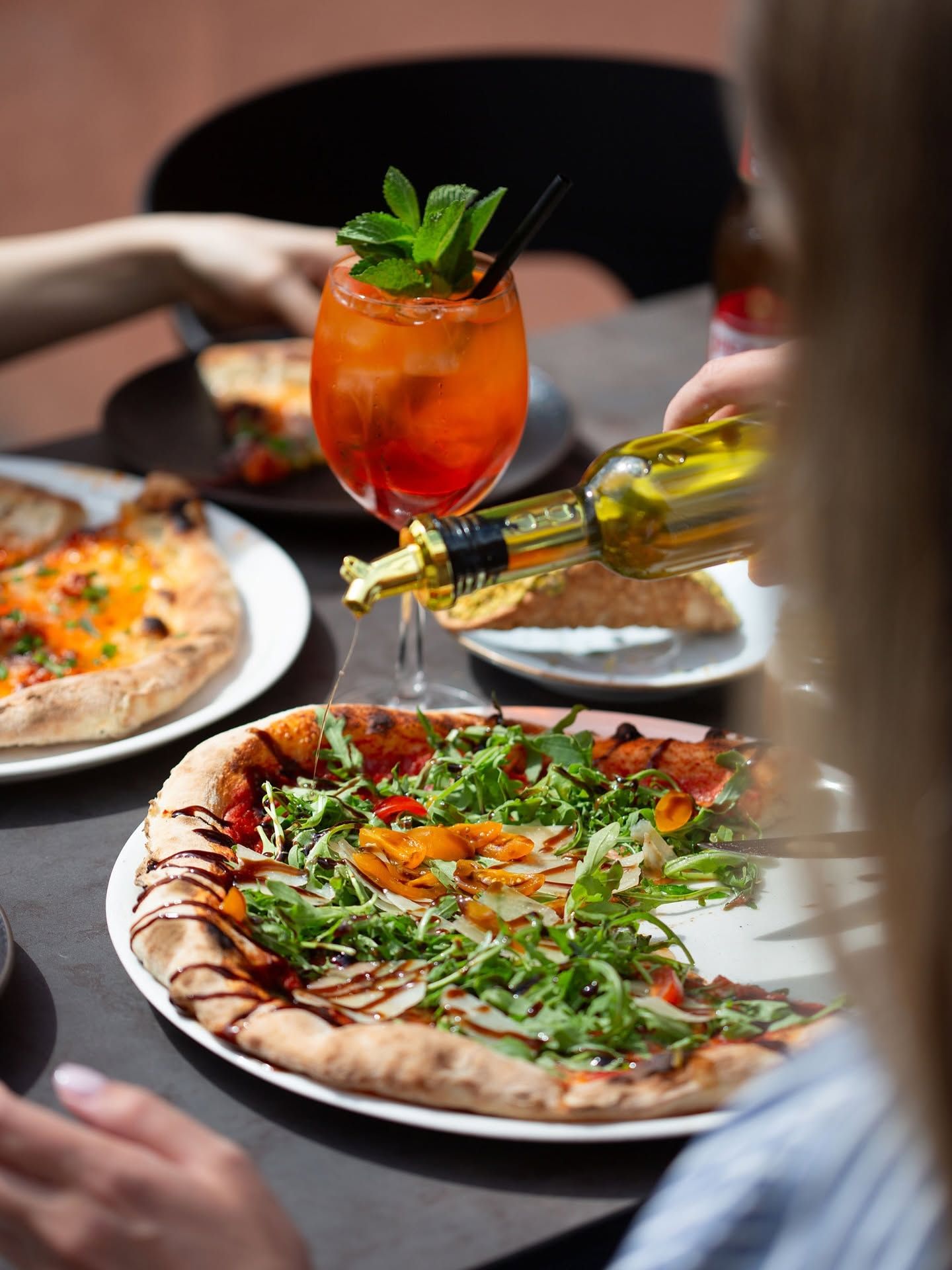 Pizza z dodatkami i drink z miętą na stole. Ktoś polewa pizzę oliwą.