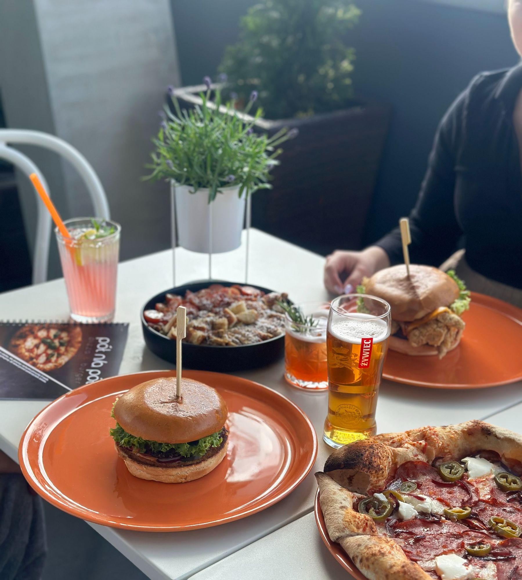 Stół z burgerami, piwem, pizzą i drinkiem, w tle roślinność i menu restauracji.