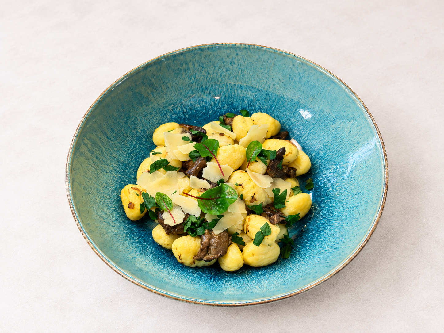 Żółte gnocchi z grzybami, plasterkami parmezanu i świeżą pietruszką na niebieskim talerzu.