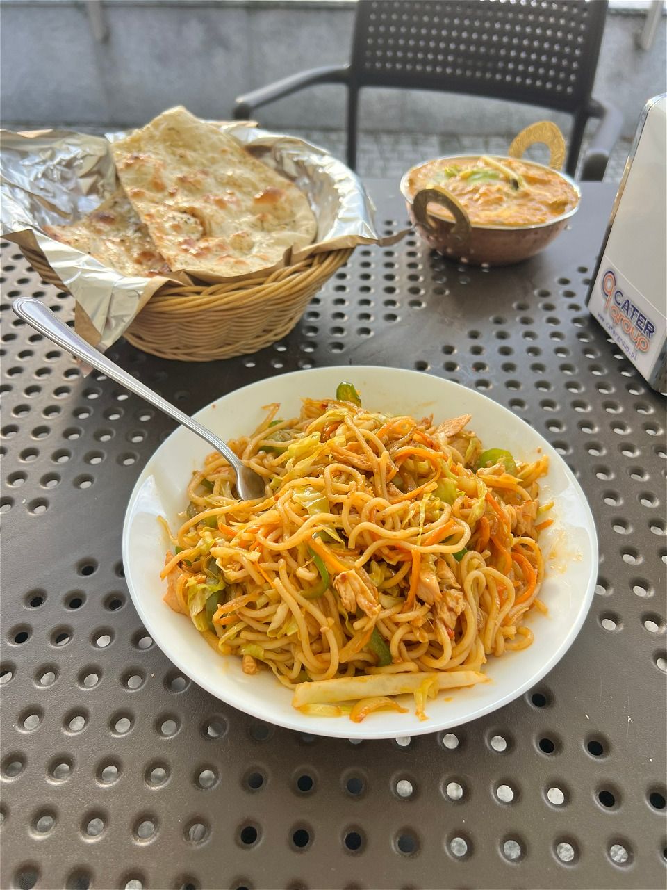 Talerz makaronu z warzywami i kurczakiem, koszyk z chlebem i miseczka z curry w tle na metalowym stole.
