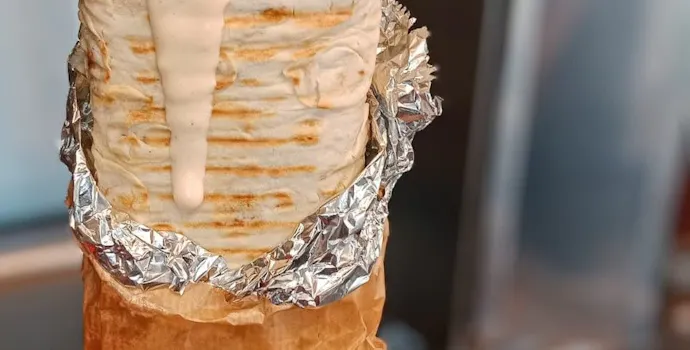 Kebab w tortilli z mięsem, warzywami i sosem, podany w papierowej torebce.