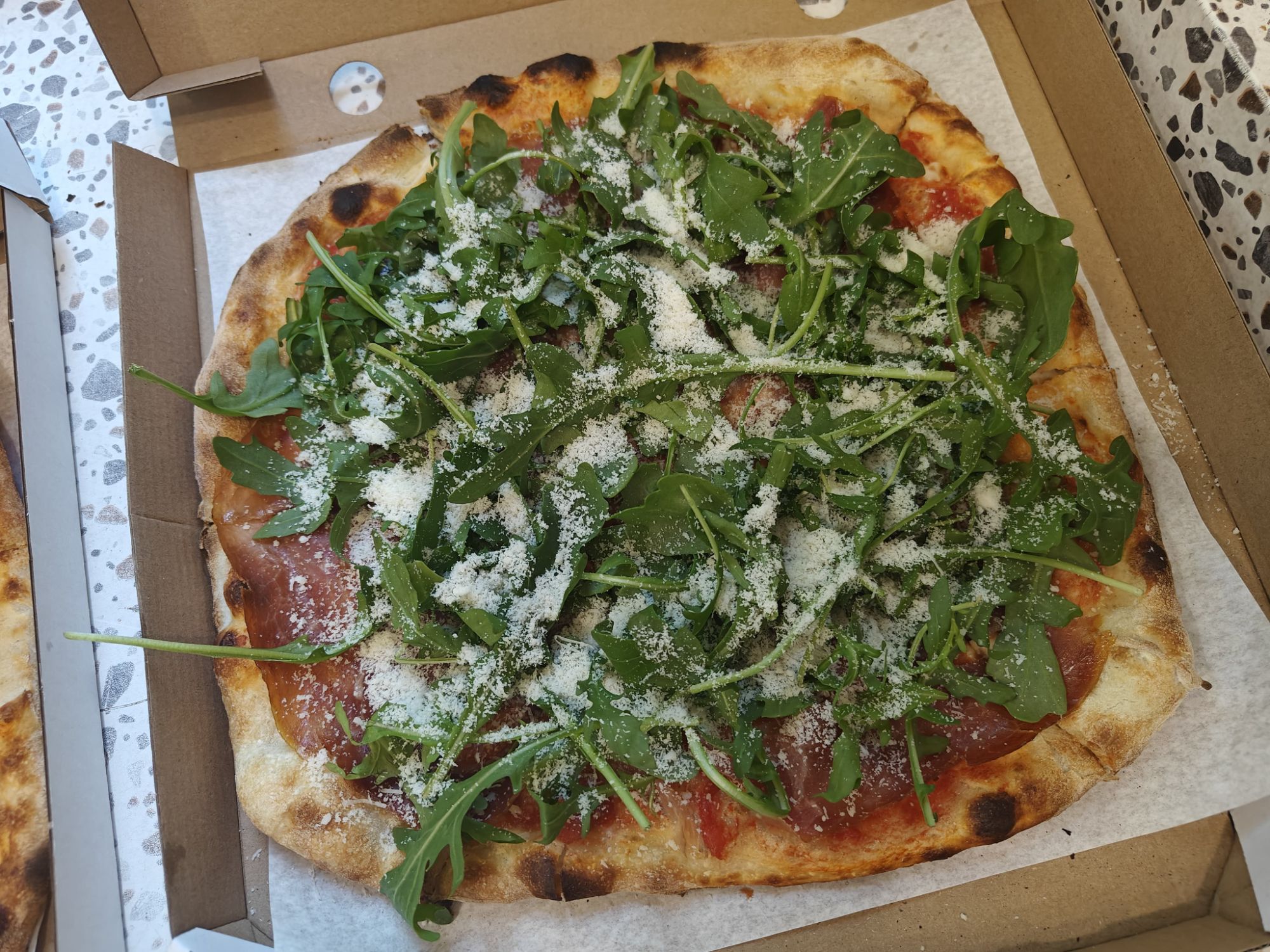 Pizza z sosem pomidorowym, serem parmezanem i świeżą rukolą na chrupiącym cieście, podana w kartonowym pudełku.