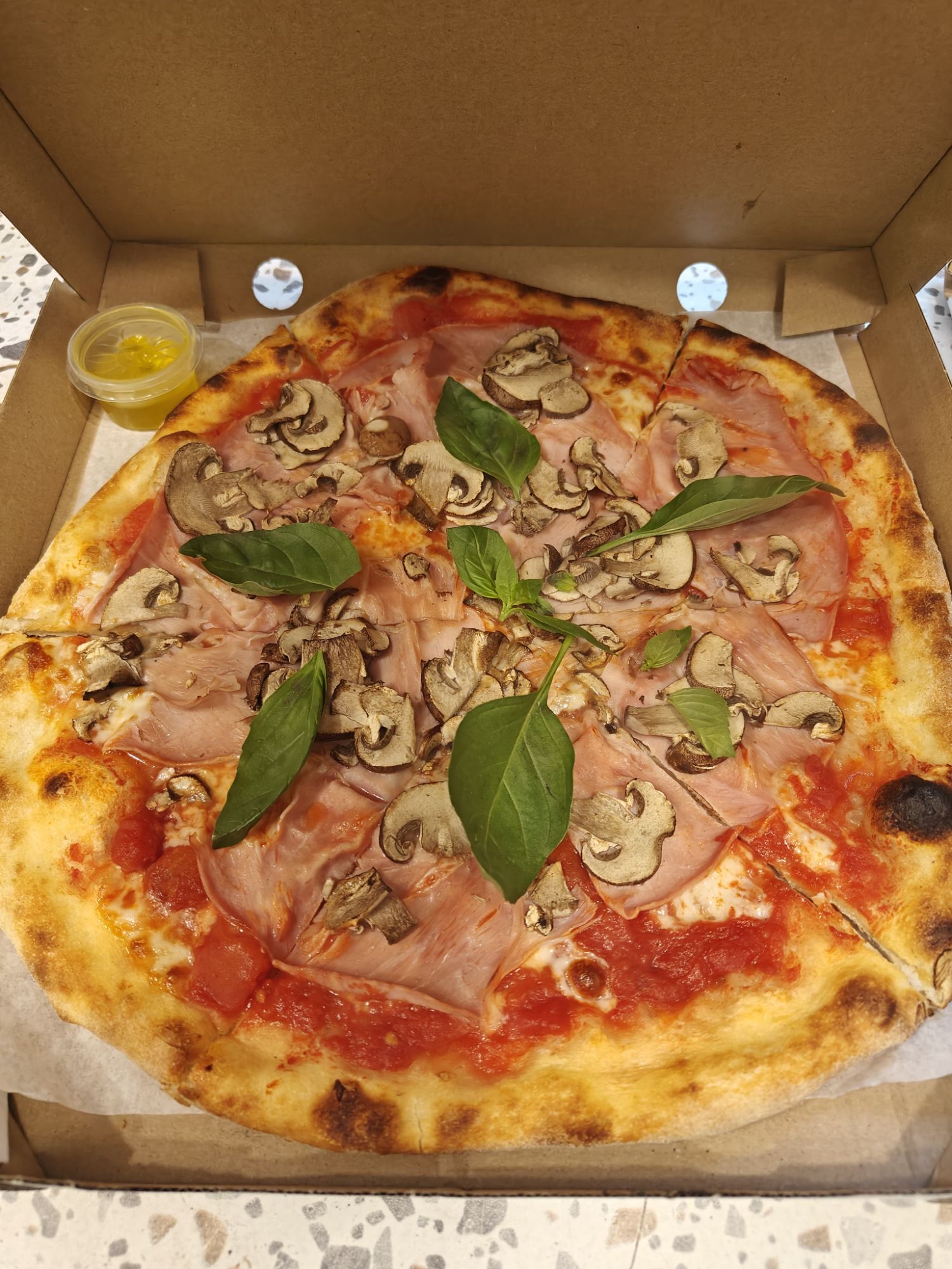 Pizza z sosem pomidorowym, szynką, pieczarkami i bazylią, podana w kartonowym pudełku.