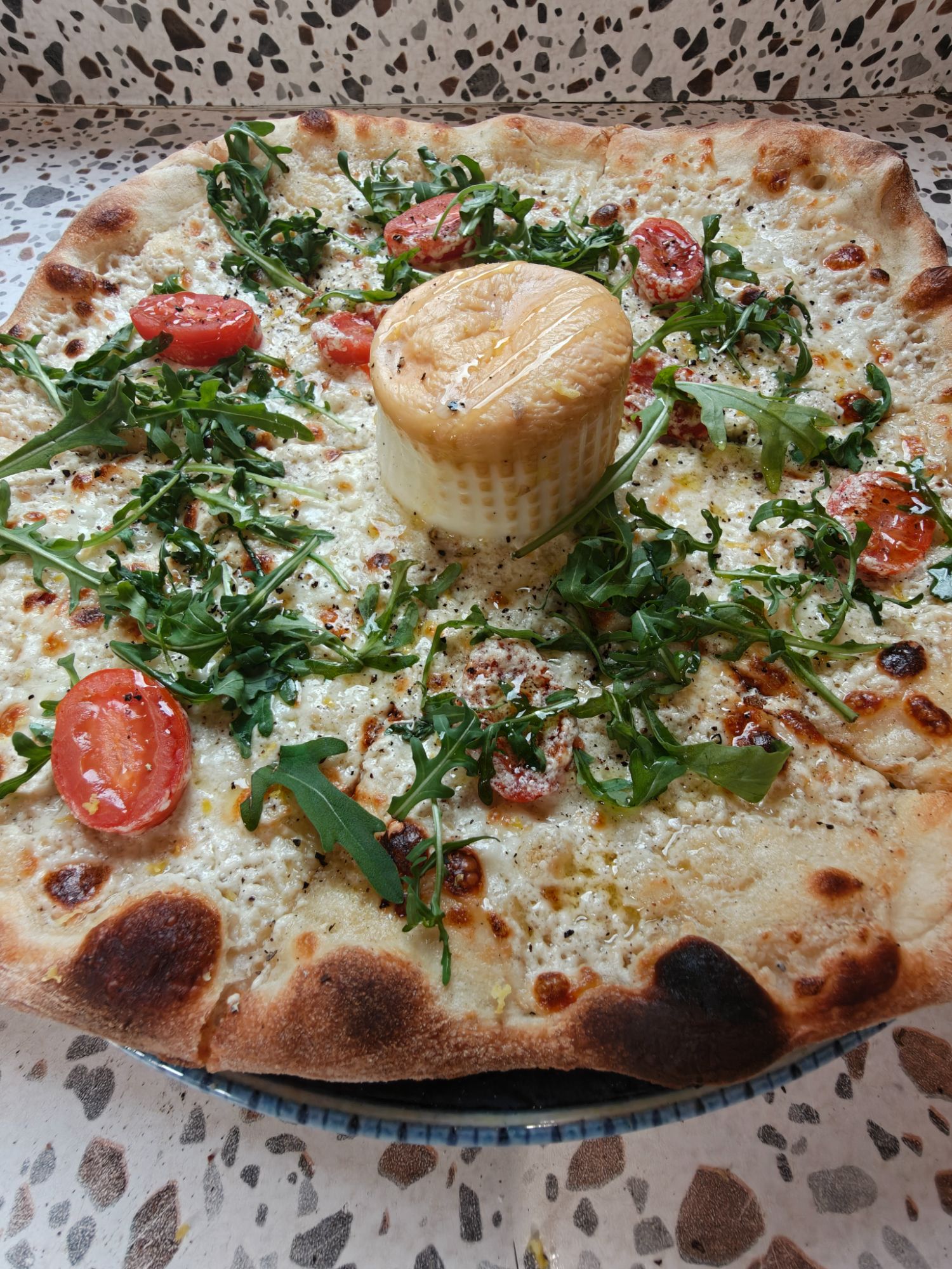 Pizza z serem, pomidorami i rukolą, z dużym serem na środku, na wzorzystym tle.