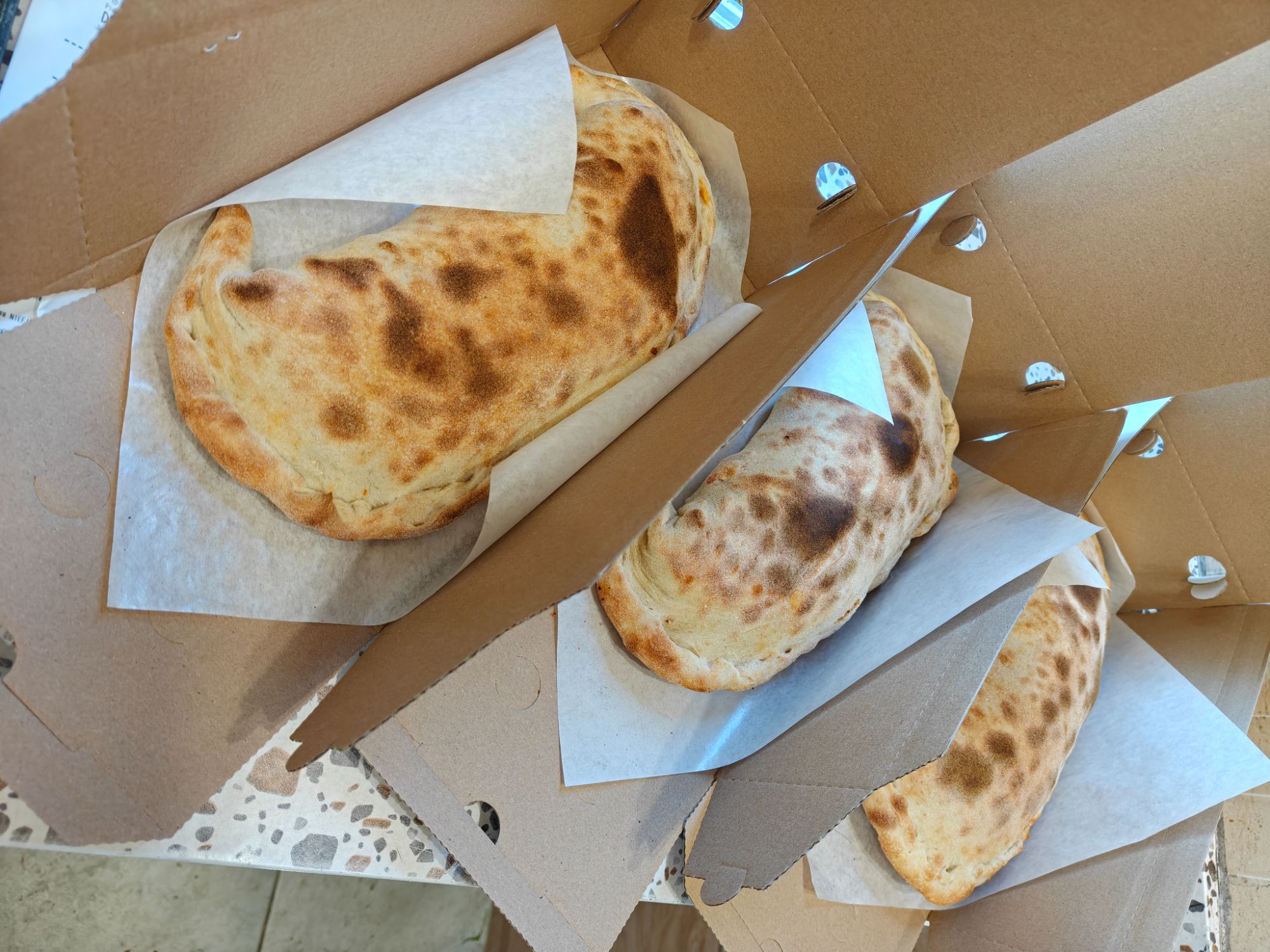 Trzy calzone w opakowaniach papierowych, leżące na blacie w jasnym pomieszczeniu.