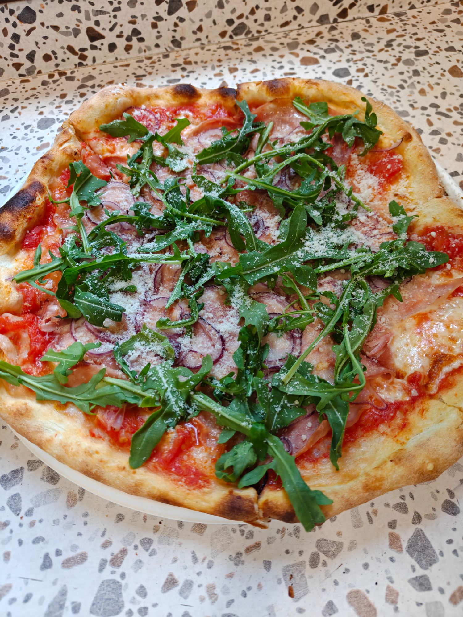 Pizza z rukolą, szynką i parmezanem na kruchym cieście, podana na blacie w drobny wzór.