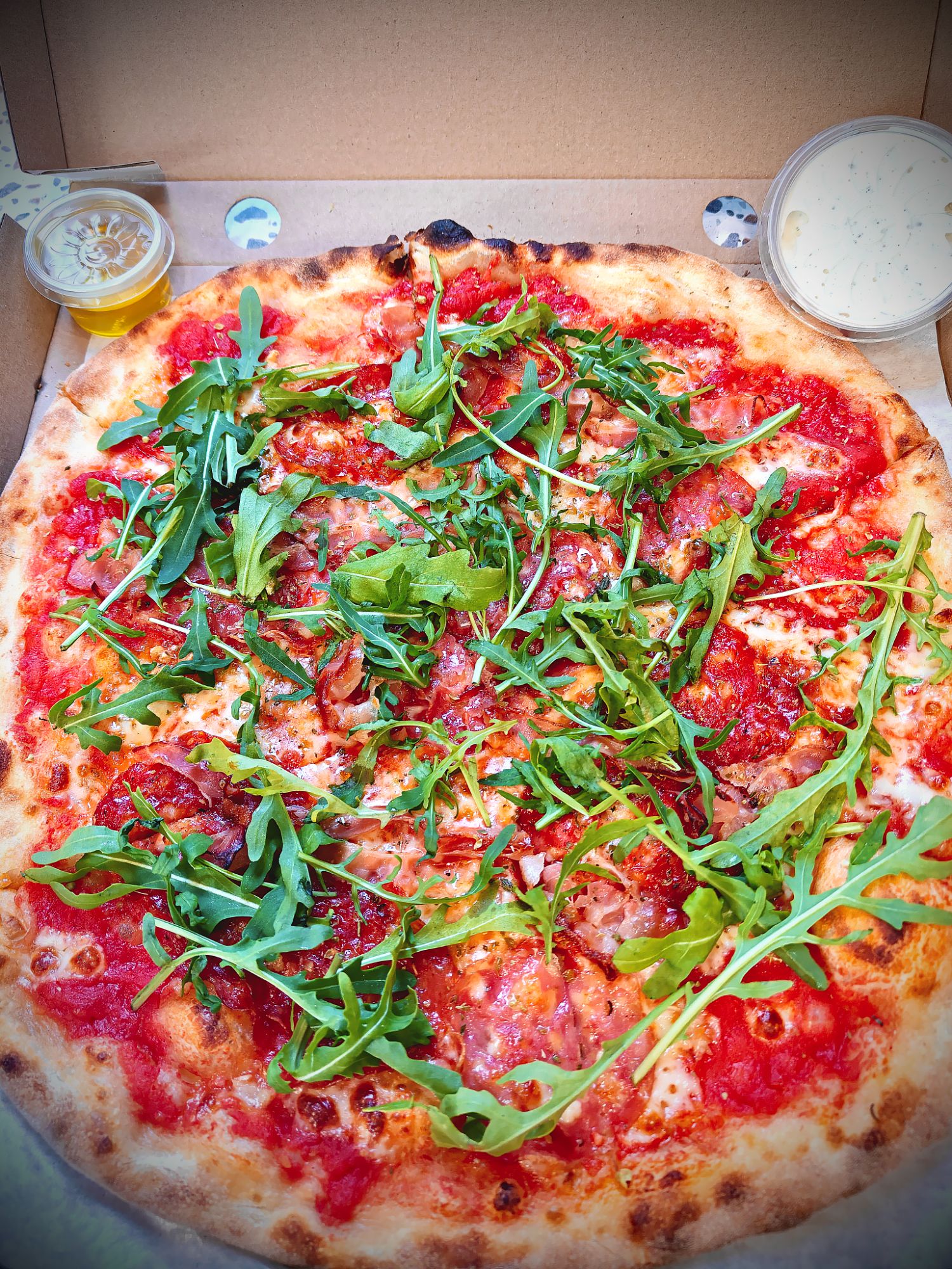 Pyszna pizza z sosem pomidorowym, serem, szynką i rukolą, podana w opakowaniu na wynos z dodatkami.
