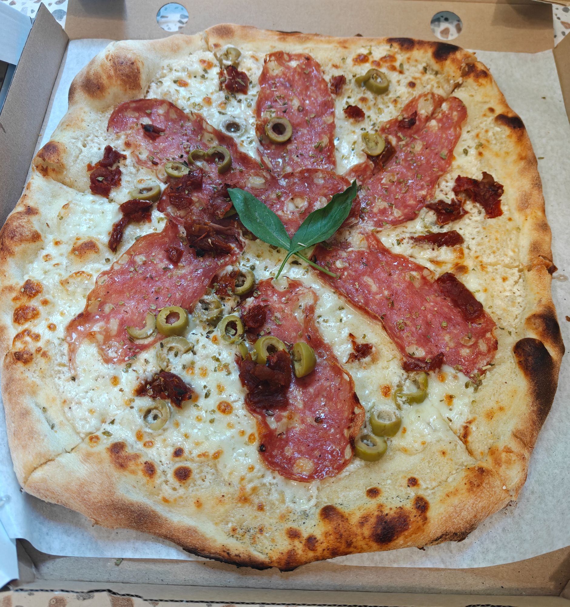 Pizza z szynką salami, oliwkami i suszonymi pomidorami, podana na wypieczonym cieście z bazylią na wierzchu.