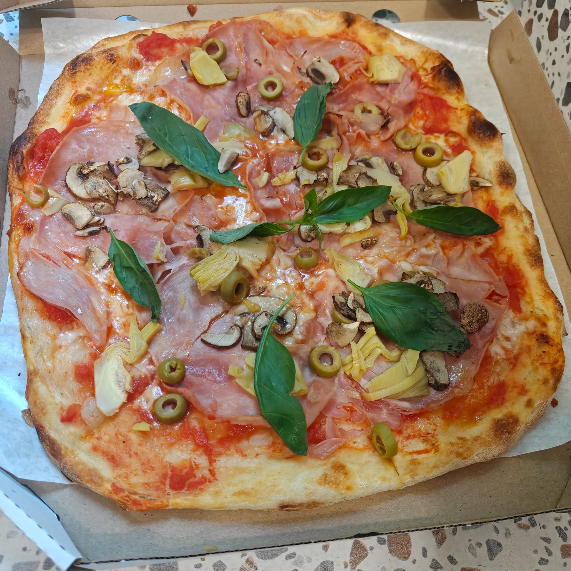 Pizza z szynką, pieczarkami, oliwkami i bazylią, podana na tekturowym opakowaniu.
