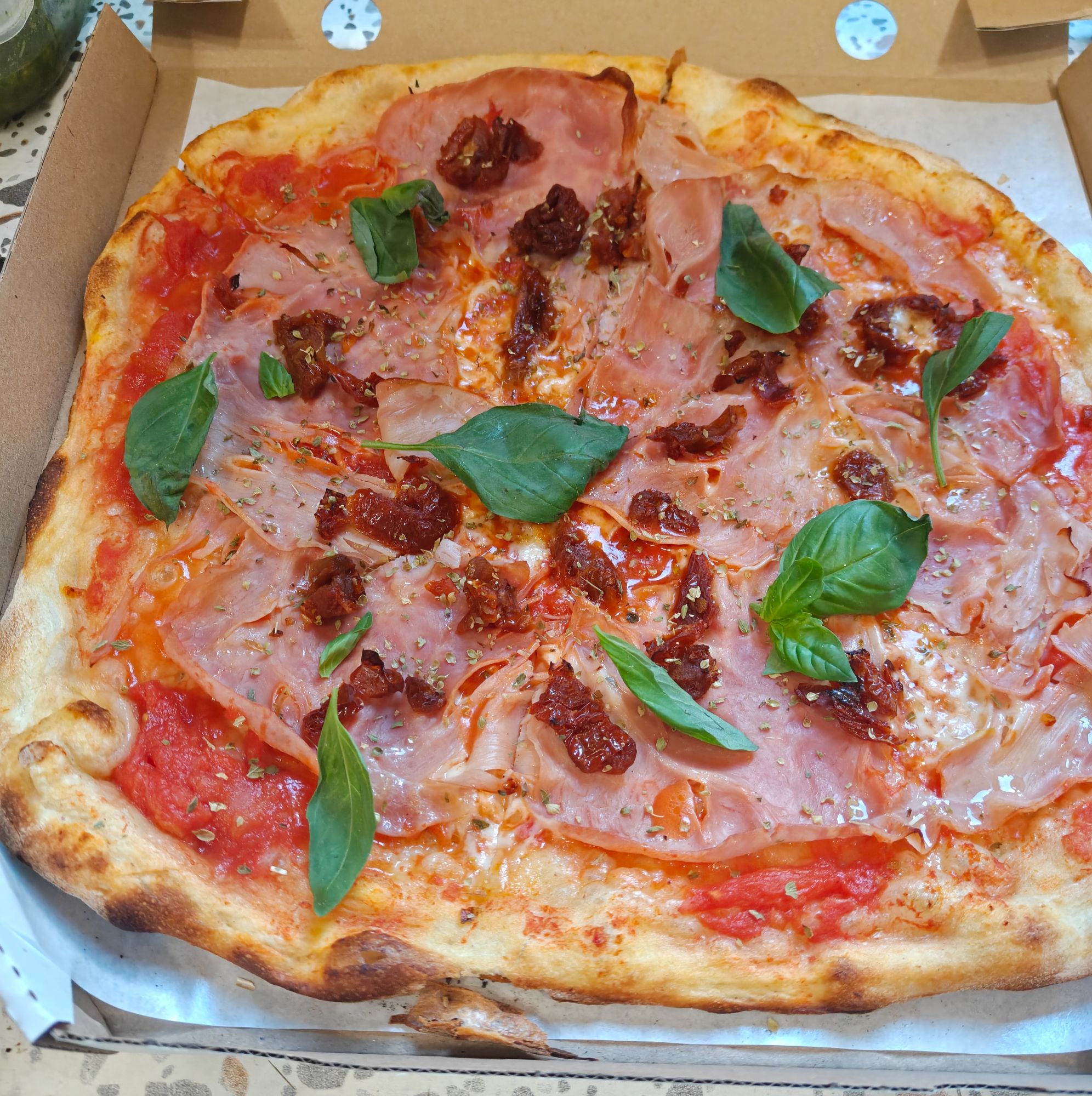 Pizza z pomidorami, szynką, suszonymi pomidorami i świeżą bazylią w kartonowym pudełku.
