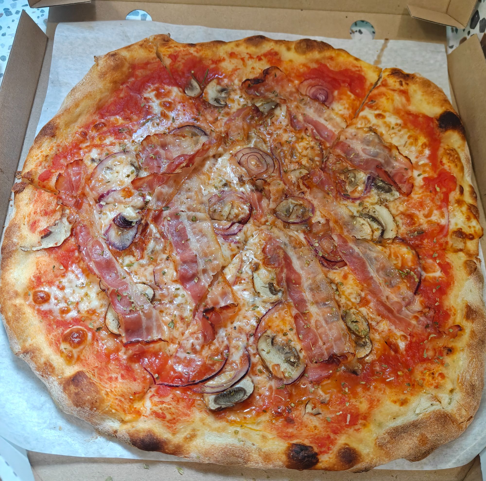 Pizza z sosem pomidorowym, serem, boczkiem, pieczarkami i cebulą, podana w kartonowym pudełku.