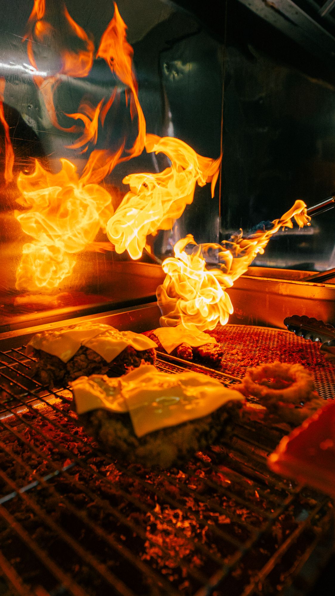 Grill z płomieniami, na którym smażą się burgery z serem.