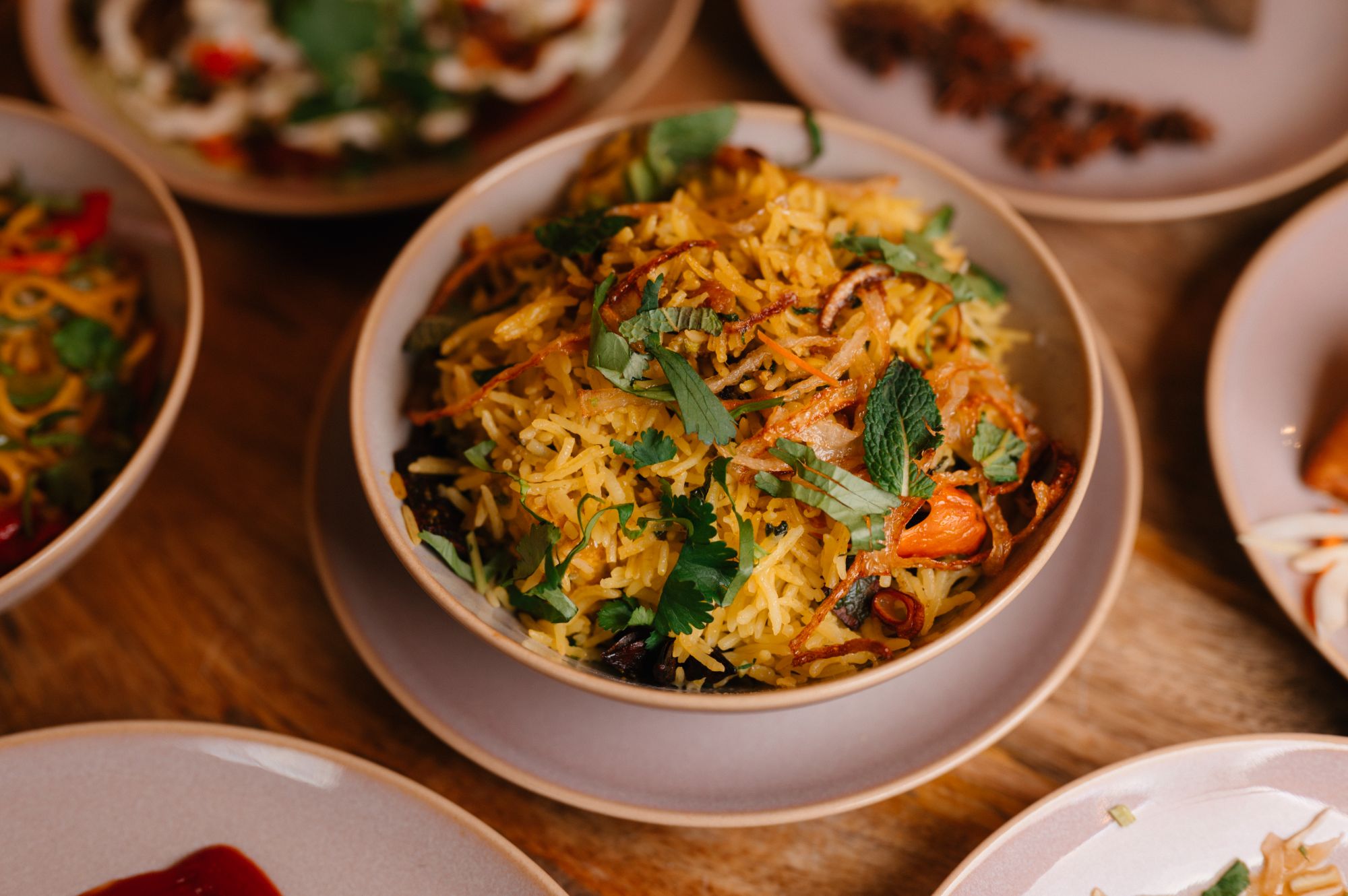 Kolorowa biryani w misce, posypana świeżą kolendrą i miętą, otoczona talerzami z różnymi potrawami.