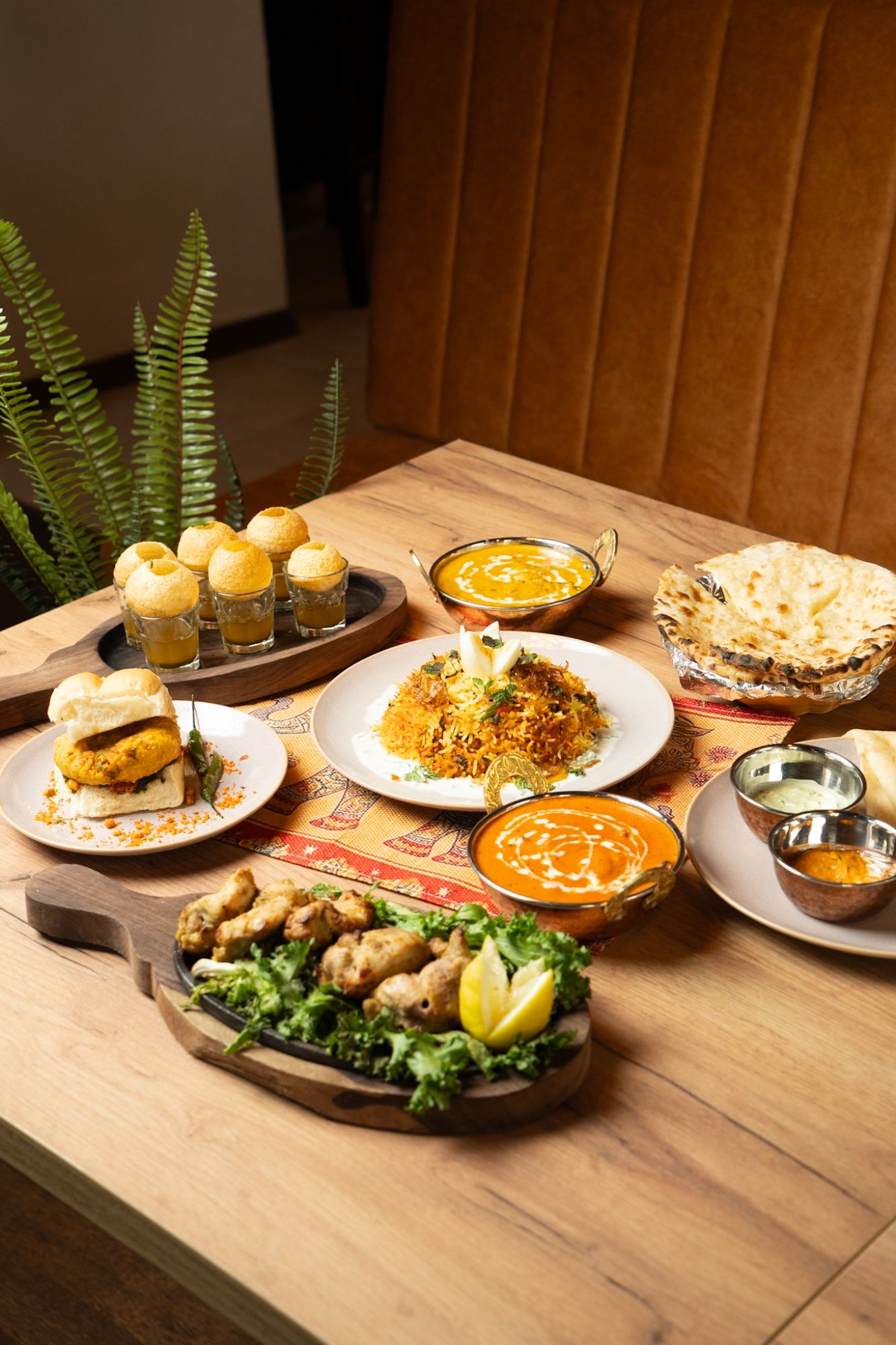 Stół z różnorodnymi potrawami: biryani, chleb, sałatki i przekąski, w otoczeniu zielonych roślin.