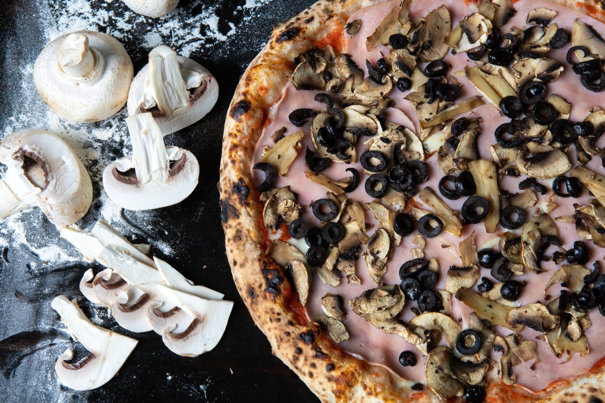 Pizza cu ciuperci și măsline negre, alături de ciuperci tăiate, pe o suprafață presărată cu făină.