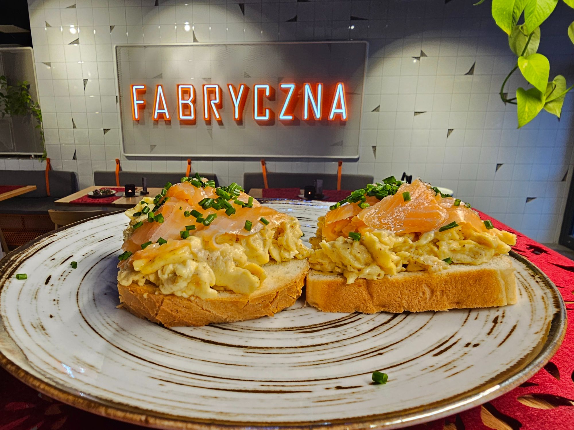 Tost z jajecznicą, wędzonym łososiem i szczypiorkiem, podany na talerzu z napisem "FABRYCZNA" w tle.