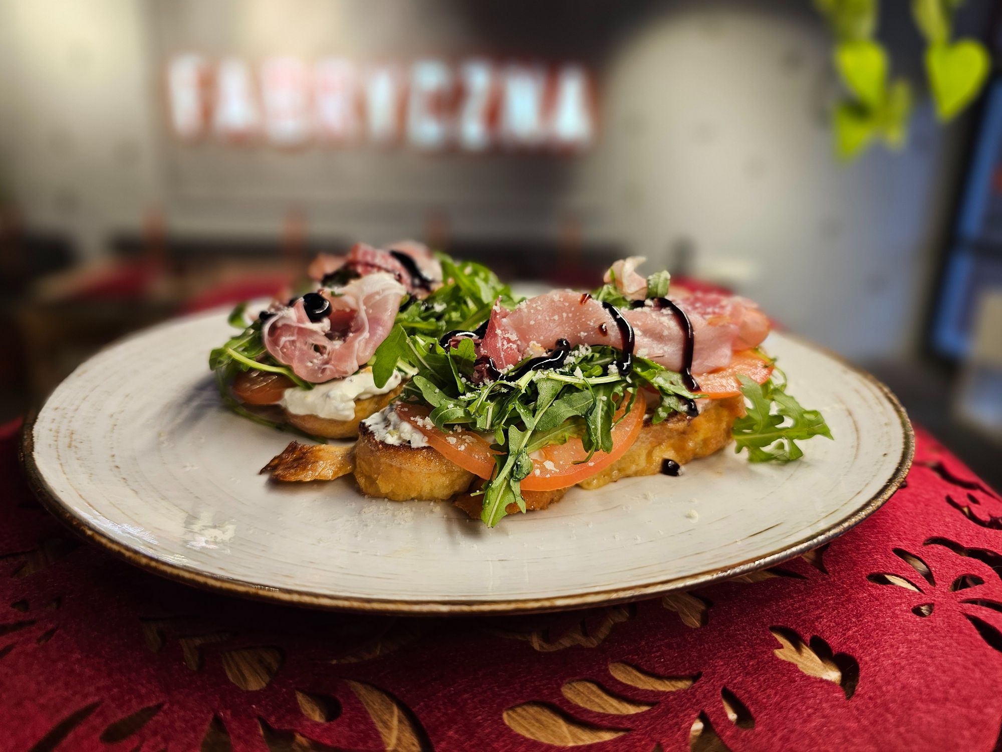 Talerz z bruschettą z pomidorami, rukolą, serem i szynką, podany na tle stylowego wnętrza restauracji.