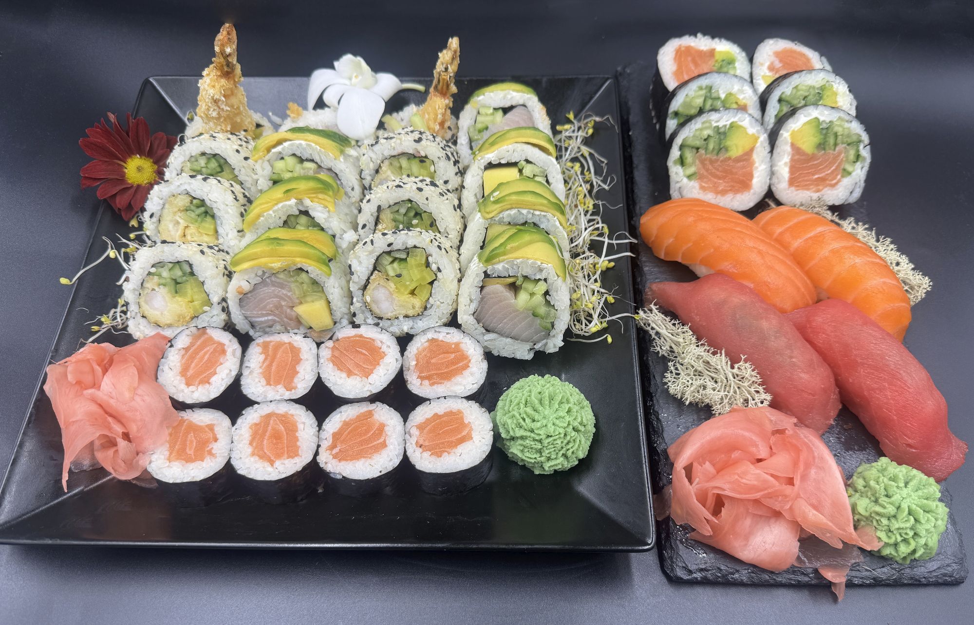 Sushi zestaw: rolki z owocami morza, łososiem, tuńczykiem i wasabi, ozdobione kwiatem i ziołami na czarnym talerzu.