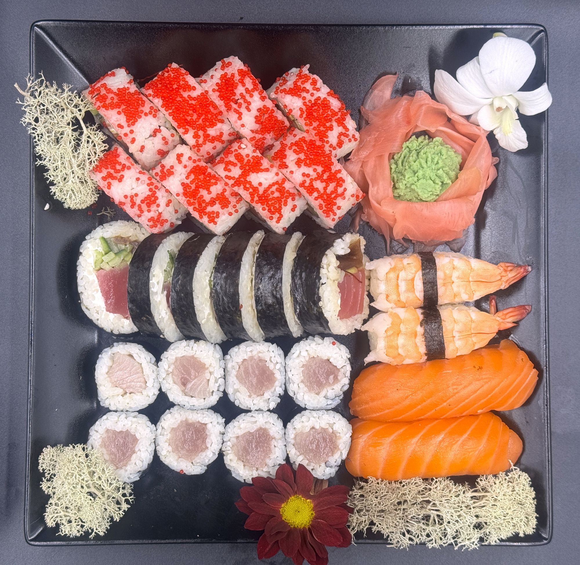 Deska sushi z różnorodnymi rolkami, krewetkami, łososiem i kwiatem ozdobnym.