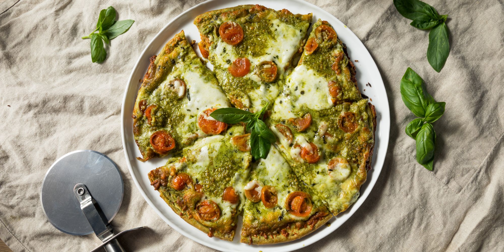 Pizza z pesto, pomidorami i serem, pokrojona na kawałki, na talerzu z bazylią w tle.