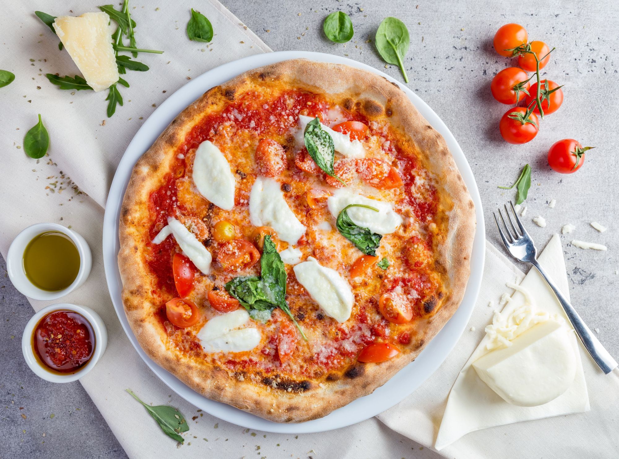 Pizza Margherita z pomidorkami, bazylią i serem mozzarella na talerzu, otoczona składnikami i dodatkami.
