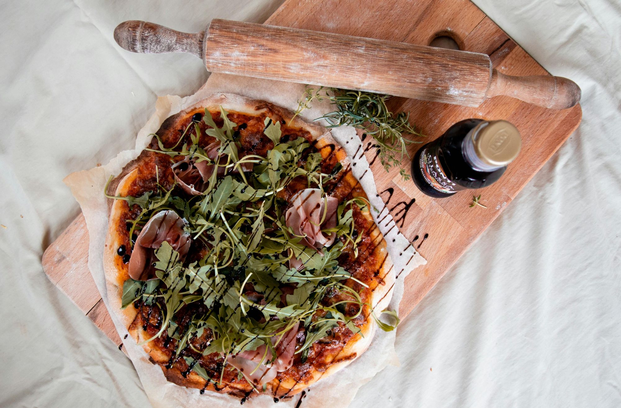 Pizza z wędliną, świeżymi ziołami na drewnianej desce, obok wałek do ciasta i sos w butelce.