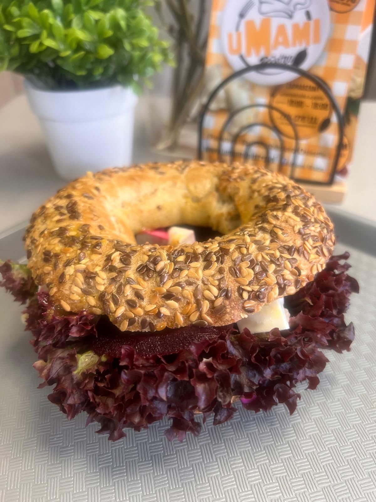 Bagel z ziarnami, sałatą, burakiem i serem, podany na szarym talerzu z rośliną w tle.