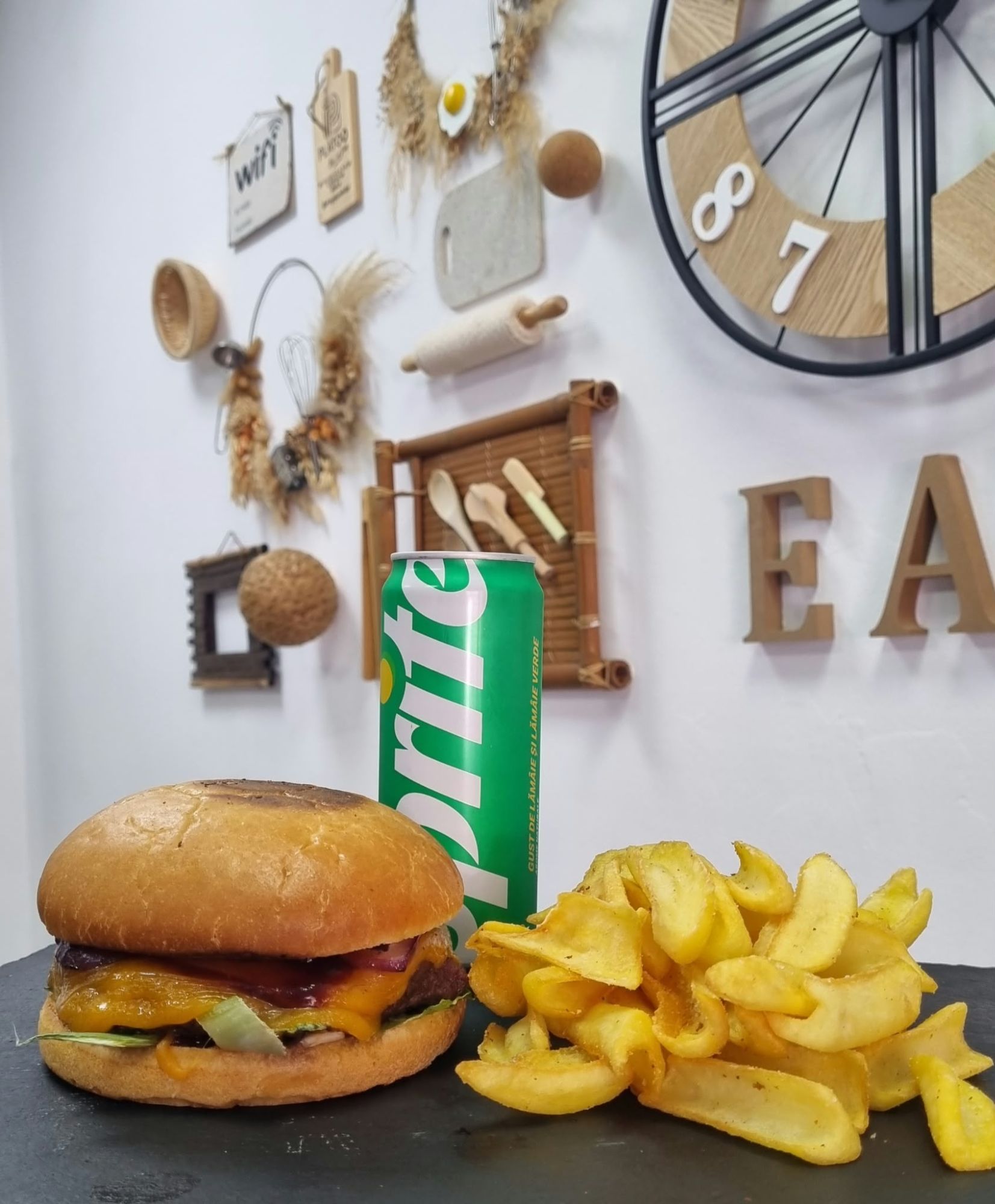 Burger cu brânză, cartofi prăjiți și o băutură Sprite, prezentate pe un fundal decorativ.