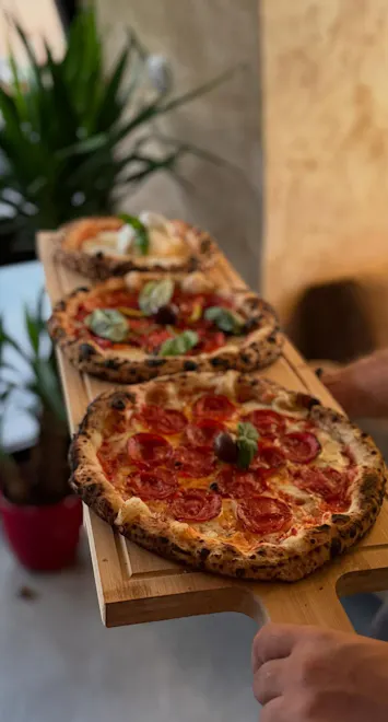 Tri pice pizze na drvenoj dasci, s rajčicama i bosiljkom, u restoranu s biljkama u pozadini.