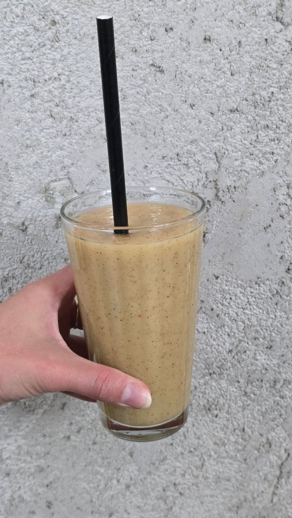 Kobieta trzyma szklankę smoothie w kolorze beżowym z czarną słomką, na tle szarej ściany.
