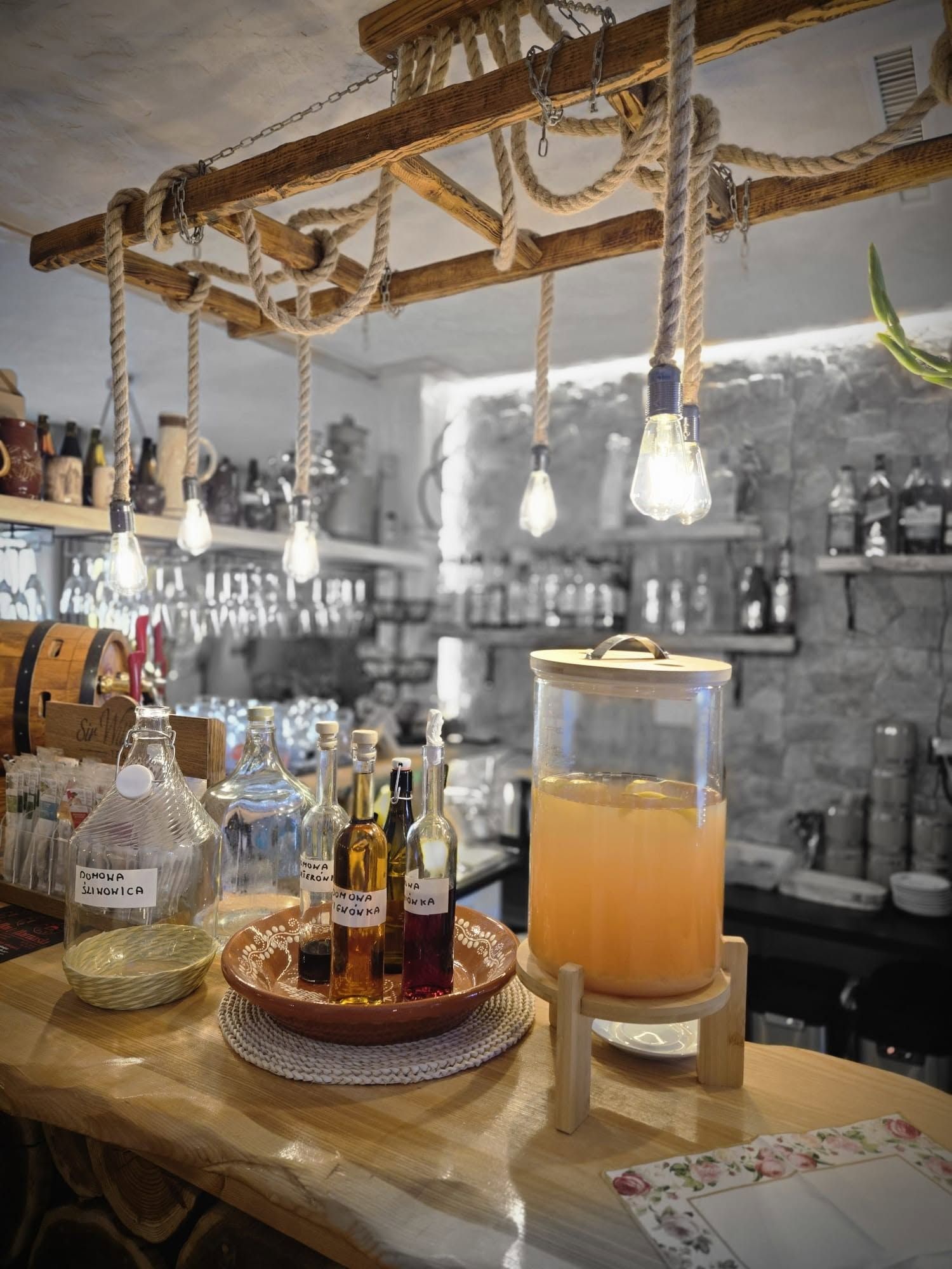 Bar z butelkami napojów i owocowym drinkiem, oświetlony ciepłym światłem wiszących lampek.