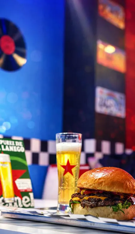 Burger z dodatkami i piwo w szklance, ustawione na stole w kolorowym retro lokalu.