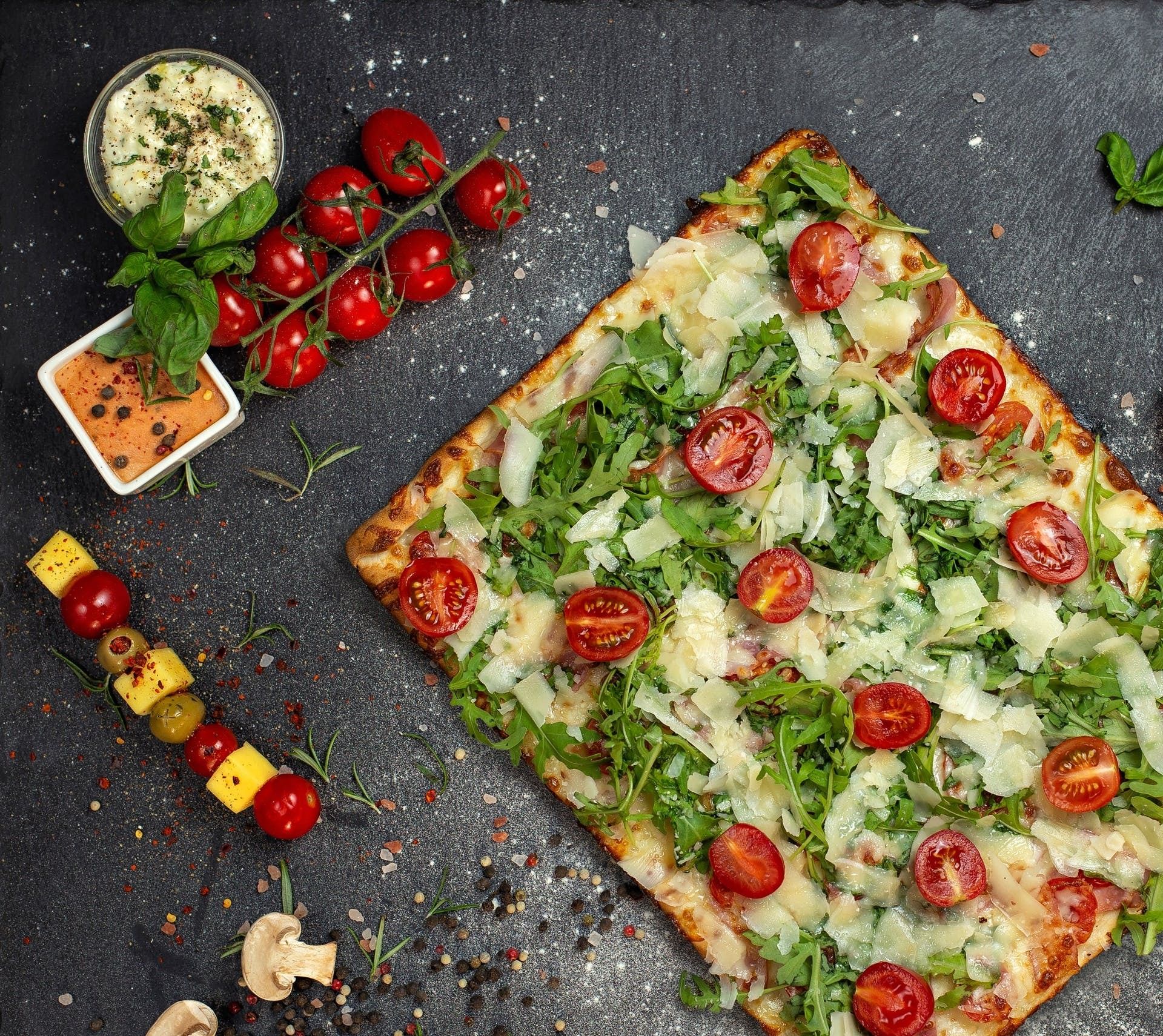 Pizza cu roșii cherry, rucola, parmezan și sosuri, pe un fundal de piatră neagră, alături de legume proaspete.